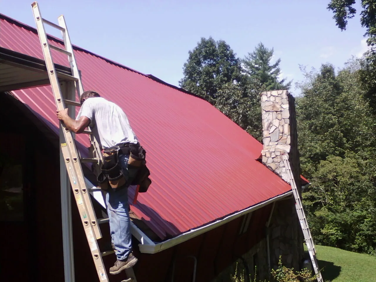 Roofer on a ladder beside a red metal roof and stone chimney — Soffit & Fascia Repair by Roofing Brothers in Grover